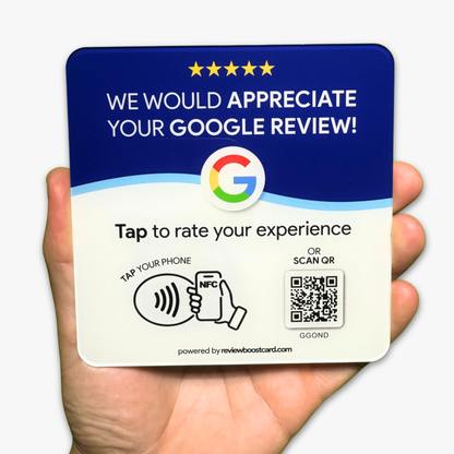 A hand holding a square card that encourages customers to leave a Google review. The card is predominantly blue and white, with a colorful Google logo in the center. It features the text, "WE WOULD APPRECIATE YOUR GOOGLE REVIEW!" followed by "Tap to rate your experience." Below this, there is an illustration of a hand holding a phone with an NFC symbol and a QR code. The card is powered by reviewboostcard.com