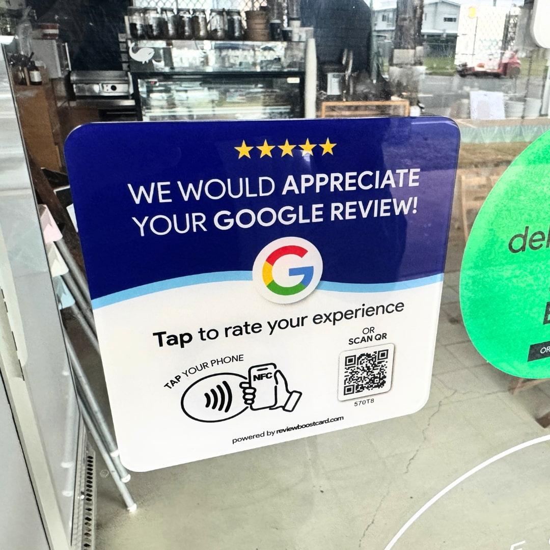 A ReviewBoost plaque displayed on a glass door of a café, encouraging Google reviews with a QR code and NFC tap feature for easy review submission
